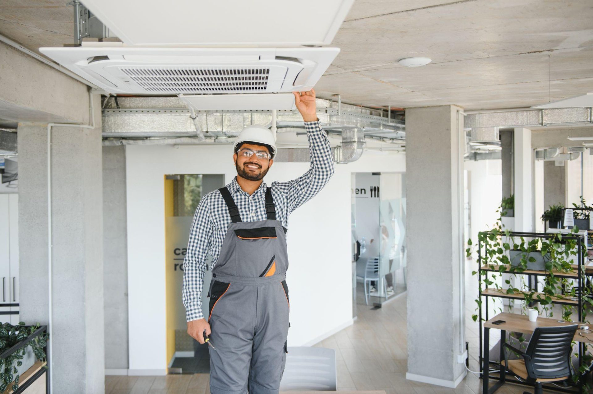 Un homme souriant, coiffé d'un casque blanc, portant des lunettes de sécurité, une chemise à carreaux et une salopette grise, se tient à l'intérieur. De la main droite, il touche un climatiseur rectangulaire blanc fixé au plafond. Il tient un outil jaune dans sa main gauche. À l'arrière-plan, on aperçoit un espace de bureaux avec des plafonds en béton apparent, des conduits de ventilation, des piliers blancs et quelques plantes.