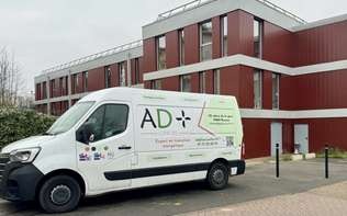 Une camionnette blanche AD+ est stationnée devant un bâtiment moderne aux façades rouges et grises. Le logo de l'entreprise, qui comprend « AD+ » en vert et noir, ainsi que du texte et un code QR, est apposé sur le côté de la camionnette.
